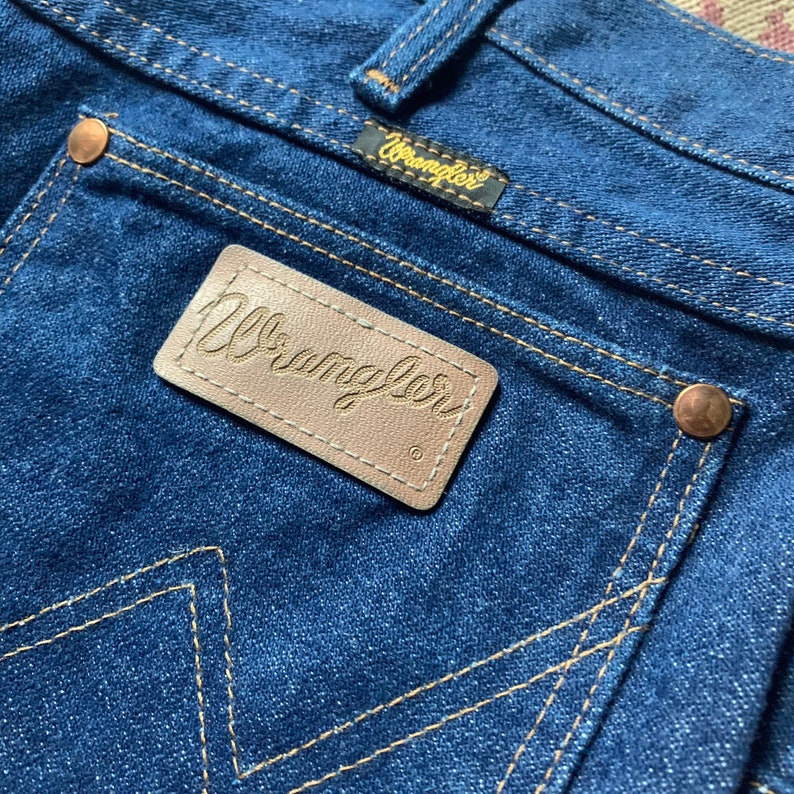 May include: Close-up of the back pocket of blue denim jeans. The pocket features a brown leather patch with the embossed word "Wrangler". The jeans have copper rivets and yellow stitching. A small tag with the "Wrangler" logo is above the pocket.