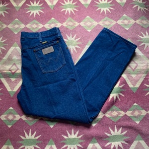 May include: Dark blue denim jeans with a classic straight leg cut. The jeans feature a brown leather patch with the Wrangler logo on the back pocket. The jeans are laid flat on a patterned rug.