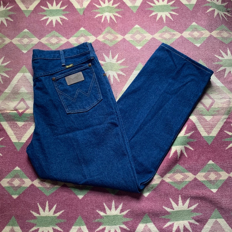 May include: Dark blue denim jeans with a classic straight leg cut. The jeans feature a brown leather patch with the Wrangler logo on the back pocket. The jeans are laid flat on a patterned rug.