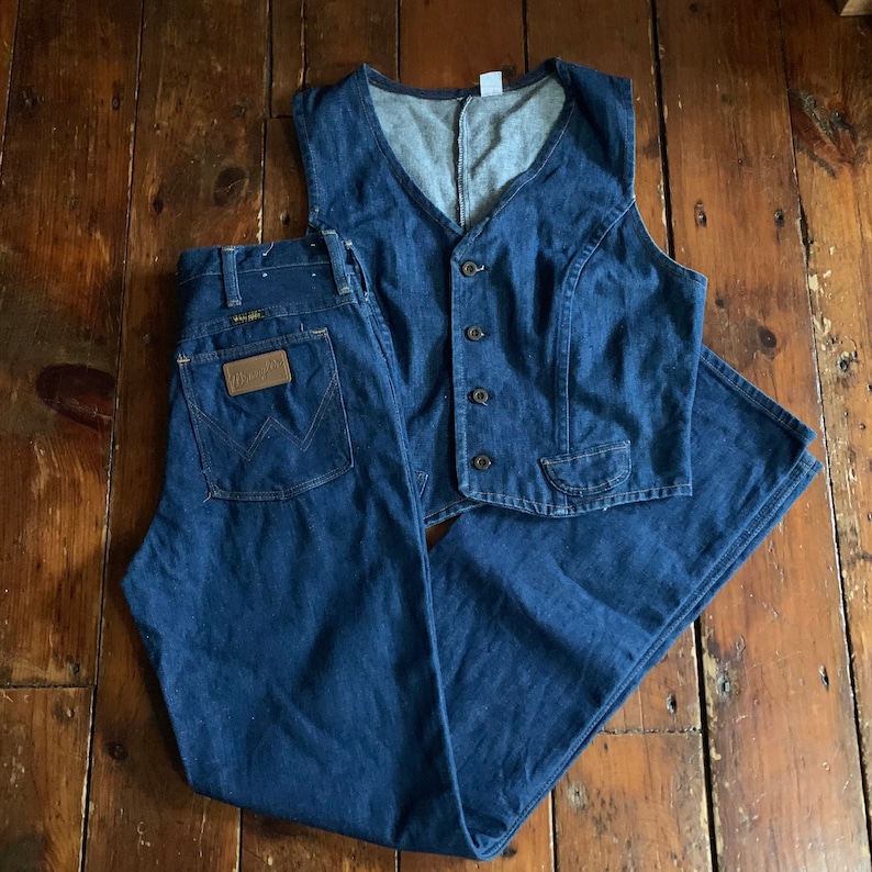 May include: A denim vest and jeans set in a medium blue wash. The vest has a button-up front and a V-neck. The jeans have a classic five-pocket design and a brown leather patch with the brand name. The set is displayed on a wooden surface.