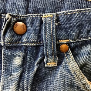 May include: Close-up of blue denim jeans with a button and a buttonhole tab. The button is bronze-colored and has the word "Wrangler" embossed on it. The jeans show signs of wear and fading.