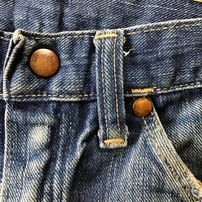May include: Close-up of blue denim jeans with a button and a buttonhole tab. The button is bronze-colored and has the word "Wrangler" embossed on it. The jeans show signs of wear and fading.