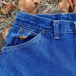 Pu&ograve; includere: Primo piano di jeans di denim blu. I jeans presentano un classico design a cinque tasche con rivetti e cuciture color rame. Un piccolo dettaglio ricamato &egrave; visibile sulla tasca anteriore. I jeans sono ambientati su uno sfondo di foglie ed erba secche.