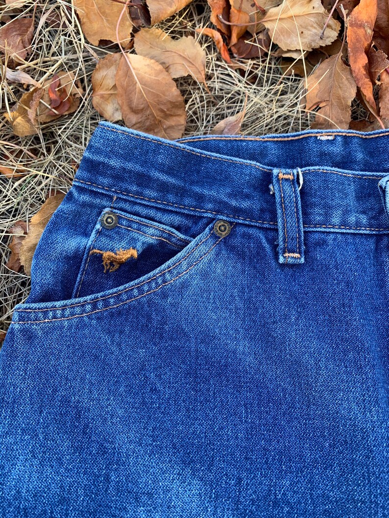 Pu&ograve; includere: Primo piano di jeans di denim blu. I jeans presentano un classico design a cinque tasche con rivetti e cuciture color rame. Un piccolo dettaglio ricamato &egrave; visibile sulla tasca anteriore. I jeans sono ambientati su uno sfondo di foglie ed erba secche.