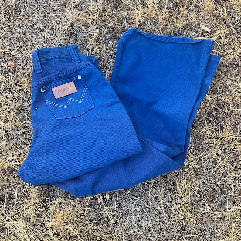 May include: A pair of blue Wrangler jeans with a brown leather patch on the back pocket. The patch features the Wrangler logo. The jeans are folded and lying on a bed of dry grass.