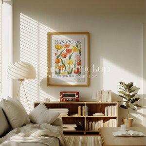 Pode incluir: Um interior de sala de estar com uma estante de madeira cheia de livros, um sof&aacute; com almofadas brancas, uma l&acirc;mpada, um p&ocirc;ster emoldurado com a palavra "HANACO" e uma ilustra&ccedil;&atilde;o floral, e um pequeno r&aacute;dio na estante.