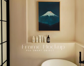 Op de afbeelding: Een ingelijste print van een blauwe berg met een witte top tegen een donkerblauwe lucht hangt aan een beige muur boven een plank met flessen en buizen met toiletartikelen. De tekst "Frame Mockup PSD Smart Object" is onder de plank gedrukt.