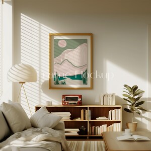 Pode incluir: Um interior de sala de estar com um sof&aacute; bege, uma estante de livros de madeira cheia de livros, um r&aacute;dio vintage, uma planta em vaso e uma impress&atilde;o de arte abstrata emoldurada em cores rosa e verde. A impress&atilde;o est&aacute; pendurada em uma parede branca com uma janela atr&aacute;s.