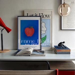 Könnte beinhalten: Ein weißer Schreibtisch mit einer roten Apfel-Illustration in einem blauen Rahmen, einem Poster mit dem Text "Let it Flow", einem Buch, einer kleinen schwarzen Figur und einer Lampe mit einem roten Schirm.