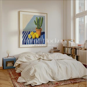 May include: A white bed with a striped comforter and a framed painting of a cactus and lemons on a blue and white striped table cloth. The painting is hanging on a white wall above the bed. There is a bookshelf with books on the right side of the image.