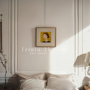 Könnte beinhalten: Ein goldfarbener Bilderrahmen mit einem Bild einer Frau mit roten Haaren, die eine schwarze Halskette trägt, hängt an einer weißen Wand über einem Bett mit weißen und beigefarbenen Kissen.