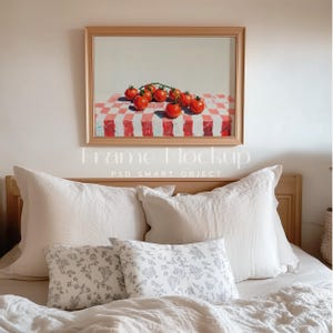 May include: A framed painting of a red checkered tablecloth with a branch of red tomatoes on top. The painting is hanging on a white wall above a bed with white linen bedding and patterned throw pillows.