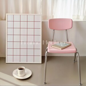 Puede incluir: Un tablero de cuadr&iacute;cula blanco enmarcado con l&iacute;neas rojas, una silla rosa con un libro en el asiento y una taza blanca con caf&eacute; en un platillo blanco.