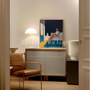 May include: A framed print of a woman sitting on a bed with a cat. The print is in a light wood frame and is hanging on a white wall. The print is above a wooden cabinet with two doors. There is a lamp on the left side of the print and a white lamp on the right side of the print.