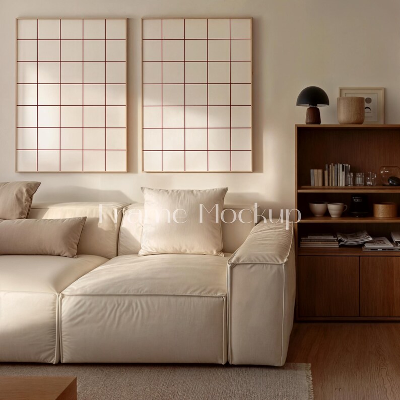 May include: Two framed wall art pieces with a grid pattern in red on a cream background. A cream-colored sectional sofa with several pillows is in front of the wall art. A wooden bookshelf and a lamp are also visible.