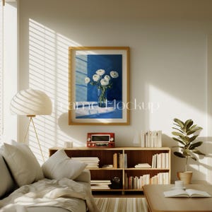 Pode incluir: Um interior de sala de estar com um sof&aacute; branco, uma estante de madeira cheia de livros, uma l&acirc;mpada e uma pintura emoldurada de flores brancas em um vaso azul sobre um fundo azul.