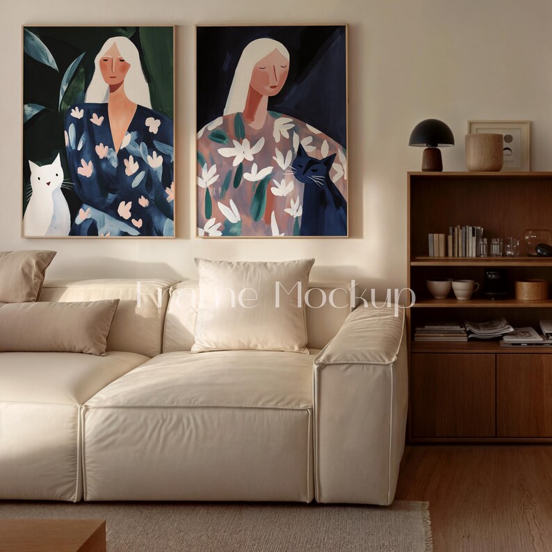 May include: Two framed art prints featuring stylized portraits of women with long blonde hair and cats. One woman wears a blue floral dress, the other a floral patterned dress. The artwork is displayed above a cream-colored sofa.