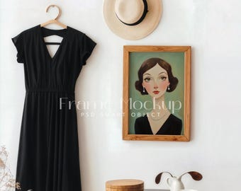 May include: A black dress with a V-neckline hangs on a wooden hanger against a white wall. A framed painting of a woman with brown hair and a black dress is on the wall. A straw hat is on the wall above the painting. A wooden box, a white pitcher, and a white bowl are on a black table.