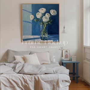 Puede incluir: Una pintura enmarcada de flores blancas en un jarrón de vidrio sobre un fondo azul. La pintura está colgada en una pared blanca sobre una cama con ropa de cama gris y blanca.