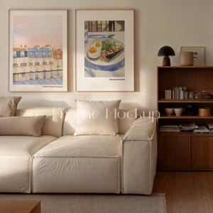 May include: Two framed watercolor prints hang above a cream-colored sofa. One print depicts a Parisian cityscape with blue and orange buildings. The other shows a plate of food with eggs and a slice of quiche. A wooden bookshelf and lamp are also visible.