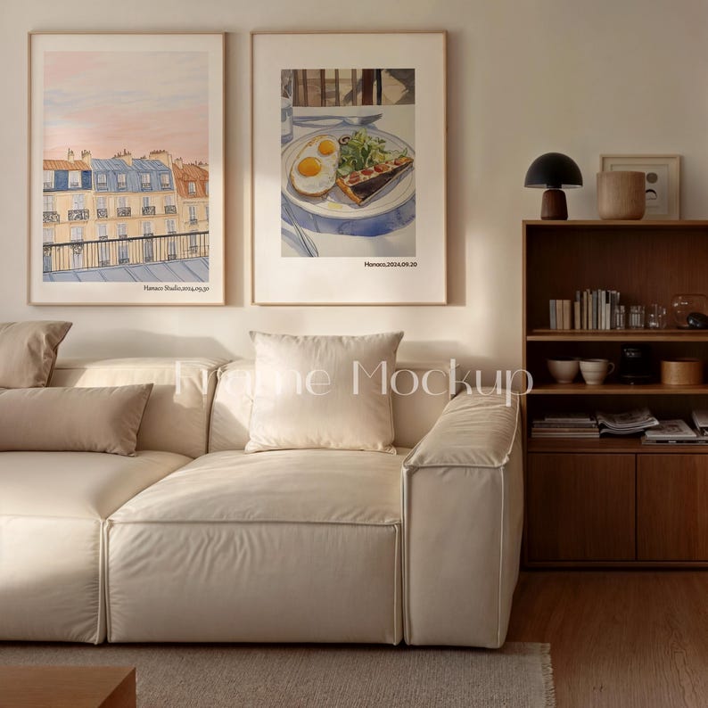 May include: Two framed watercolor prints hang above a cream-colored sofa. One print depicts a Parisian cityscape with blue and orange buildings. The other shows a plate of food with eggs and a slice of quiche. A wooden bookshelf and lamp are also visible.