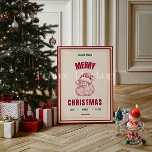K&ouml;nnte beinhalten: Ein gerahmter Weihnachtsdruck mit einer roten und wei&szlig;en Illustration des Weihnachtsmanns. Der Druck lautet "Frohe Weihnachten" und "Hanako Studio" in gr&uuml;nem Text. Der Rahmen ist rot und wei&szlig; und steht auf einem Holzboden vor einem Weihnachtsbaum.
