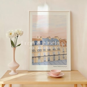 Puede incluir: Una ilustración enmarcada de una fila de edificios con un cielo rosa y azul de fondo. La ilustración está en un marco blanco y está sobre una mesa de madera marrón claro. Hay flores blancas en un jarrón sobre la mesa.