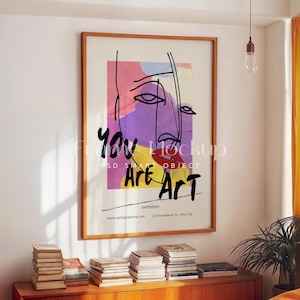 Könnte beinhalten: Ein gerahmter Kunstdruck mit einem minimalistischen Gesichtsdesign und den Worten "You Are Art" in Schwarz. Das Kunstwerk zeigt einen farbenfrohen Hintergrund mit rosa, lila und gelben Farbtönen. Der Rahmen ist warmbraun.