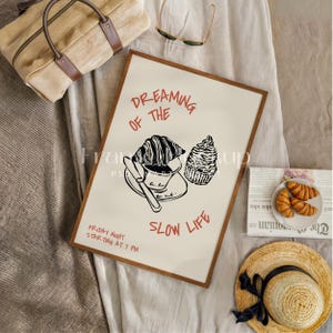 May include: A framed print with the text "Dreaming of the Slow Life" and illustrations of a croissant, coffee, and a pastry. The print is in a wooden frame, with a straw hat and a bag in the background.