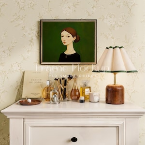 May include: A white dresser with a framed portrait of a woman with dark hair and a green background. On the dresser are a book, a lamp with a white shade, perfume bottles, makeup brushes, and a candle.