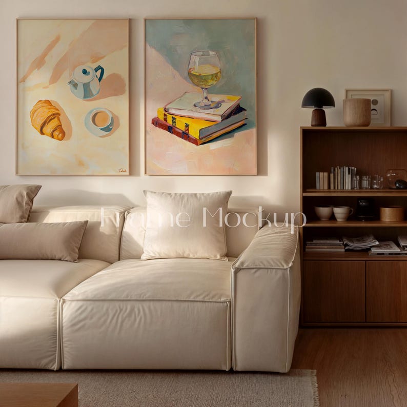 May include: Two framed paintings depict a still life. One shows a croissant, teapot, and teacup. The other features a wine glass on books. A cream-colored sofa with pillows sits below the artwork, with a wooden bookshelf and lamp in the background.