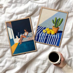 Op de afbeelding: Twee ingelijste kunstdrukken met houten frames. Eén toont een vrouw met een kat, de andere een cactus met citroenen. Een hand houdt een koffiekopje op een wit laken.