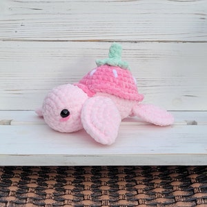 May include: A pink crocheted turtle with a strawberry on its back. The turtle has a green stem and white seeds on the strawberry. The turtle is sitting on a white wooden surface.