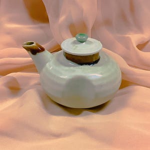 Puede incluir: Una pequeña tetera redonda de cerámica con un esmalte verde claro. El pico y el borde de la tapa son marrones. La tapa tiene un pequeño pomo verde. La tetera está sobre una tela color melocotón.