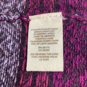 May include: A close-up of a beige fabric tag with black text listing the fabric content of a garment. The tag reads: 31% Acrylic/Acrylique, 28% Nylon/Nylon, 21% Merino Wool/Laine de Merinos, 13% Wool/Laine, 7% Angora Rabbit Hair/Poil de Lapw Angora, RN 66170, CA 32054, See Reverse for Care, Voir L'Inverse Pour Le Soin.