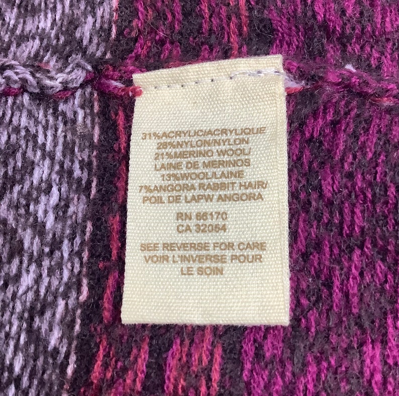 May include: A close-up of a beige fabric tag with black text listing the fabric content of a garment. The tag reads: 31% Acrylic/Acrylique, 28% Nylon/Nylon, 21% Merino Wool/Laine de Merinos, 13% Wool/Laine, 7% Angora Rabbit Hair/Poil de Lapw Angora, RN 66170, CA 32054, See Reverse for Care, Voir L'Inverse Pour Le Soin.