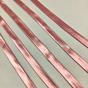 May include: Close-up of several lengths of pink satin ribbon. The ribbons are arranged diagonally across the frame, creating a pattern. The satin has a slight sheen, reflecting light. The ribbons are a soft, muted pink.