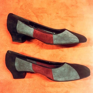May include: A pair of low-heeled shoes with a color-block design. The shoes feature black, gray, and burgundy suede panels. The shoes have a rounded toe and a small heel. The shoes are displayed on an orange background.