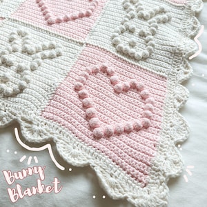 May include: A white and pink crocheted baby blanket with a scalloped edge and a heart pattern. The blanket has a "Bunny Blanket" text label.