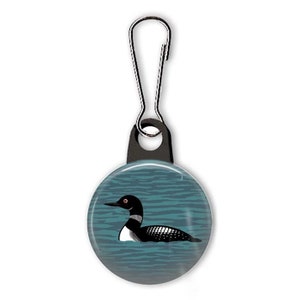 May include: A black and white loon with red eyes is swimming in blue water. The loon is on a round button attached to a black zipper pull.