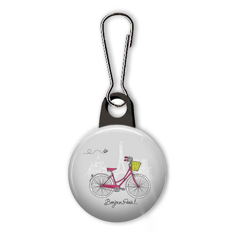 Peut inclure: Un tire-zip blanc avec un bouton rond repr&eacute;sentant un v&eacute;lo rose avec un panier jaune devant une illustration grise et blanche de la tour Eiffel et d'autres monuments parisiens. Le texte "Bonjour Paris!" est imprim&eacute; sous le v&eacute;lo.