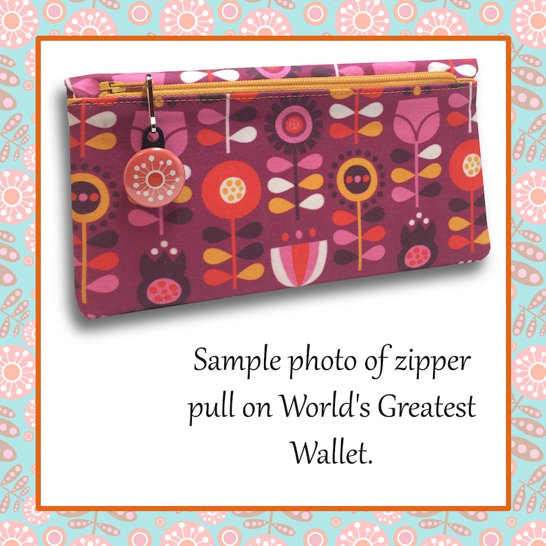 May include: A close-up of a zipper pull on a pink and red floral patterned wallet. The zipper pull is a round, red button with a white design. The text "Sample photo of zipper pull on World's Greatest Wallet." is below the wallet.