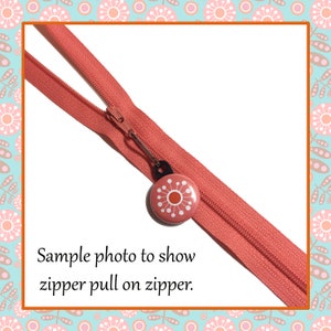 May include: A close-up of a coral colored zipper with a pink and orange flower zipper pull. The zipper pull is attached to the zipper with a silver metal ring. The text "Sample photo to show zipper pull on zipper." is below the zipper.