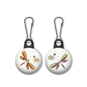 May include: Two zipper pulls with round buttons featuring a dragonfly design. The left button has an orange and brown dragonfly with a green flower. The right button has a purple and green dragonfly with a green flower. Both buttons have a white background with green polka dots.