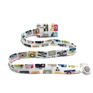 Peut inclure: Lanière en tissu blanc avec une fermeture à pression en plastique blanc. La lanière présente un motif répétitif de caméras de dessin animé colorées. La lanière a une petite pochette en tissu avec le texte "TinyTote" imprimé dessus.