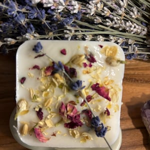 May include: A white square soap bar with dried lavender, rose petals, and jasmine flowers embedded in it. The soap is sitting on a white ceramic dish.