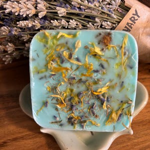 May include: A light blue soap bar with dried lavender and yellow flowers embedded in it. The soap is sitting on a light blue ceramic soap dish.