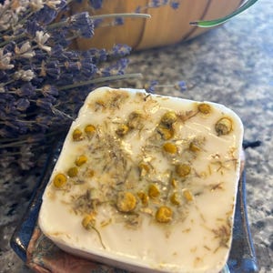 May include: A square, off-white bar of soap with dried chamomile flowers and herbs embedded on top. The soap rests on a small, blue ceramic dish with a patterned fabric underneath. A bundle of dried lavender is in the background.