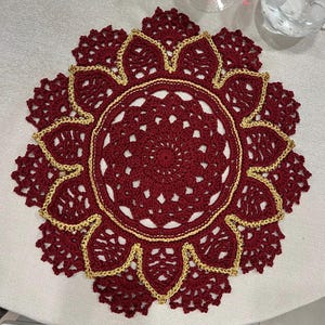 May include: A burgundy crochet doily with a gold trim. The doily has a circular shape and a delicate, intricate design.