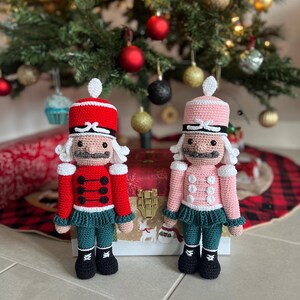 May include: Two handmade crochet nutcracker dolls. One is red with black buttons and green pants, the other is pink with white buttons and green pants. Both have white hair and hats, and are standing in front of a Christmas tree.